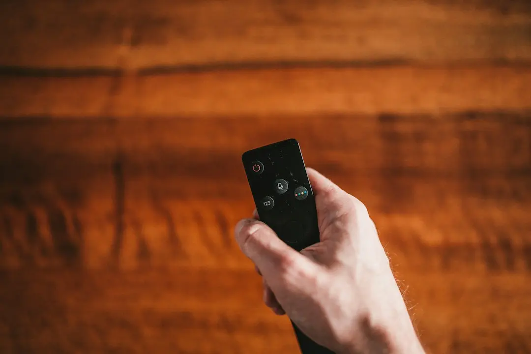 A hand holding a TV remote over a wooden table, as when choosing a player app for MeezzyTV